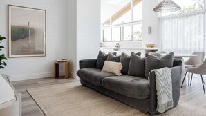 A modern living room with a grey sofa, a small wooden side table, and a large window with sheer curtains.