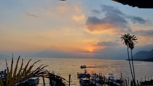 A sunset view over a calm sea with boats docked at a pier, a mountain in the background, and a palm tree on the right. - Hotel Tzanjuyu Bay (Panajachel)