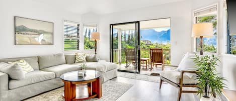 Un salón amplio con un sofá, una mesa de centro y una silla. Hay ventanas grandes con vistas a zonas verdes y montañas.