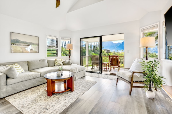 Un salon spacieux comprenant un canapé, une table basse et un fauteuil. De grandes fenêtres offrent une vue sur la verdure et les montagnes.