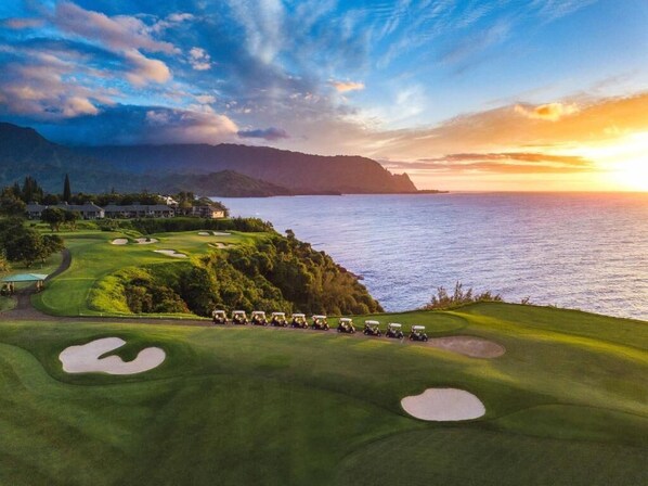 Campo de golf con búnkeres y una fila de carritos de golf, con vistas a un cuerpo de agua y montañas al atardecer.