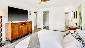 A hotel room with a large bed, a wooden cabinet with a flat-screen TV, a ceiling fan, and a door leading to another room. - Private Princeville Retreat w/ Hanalei Bay View!
 (Princeville)