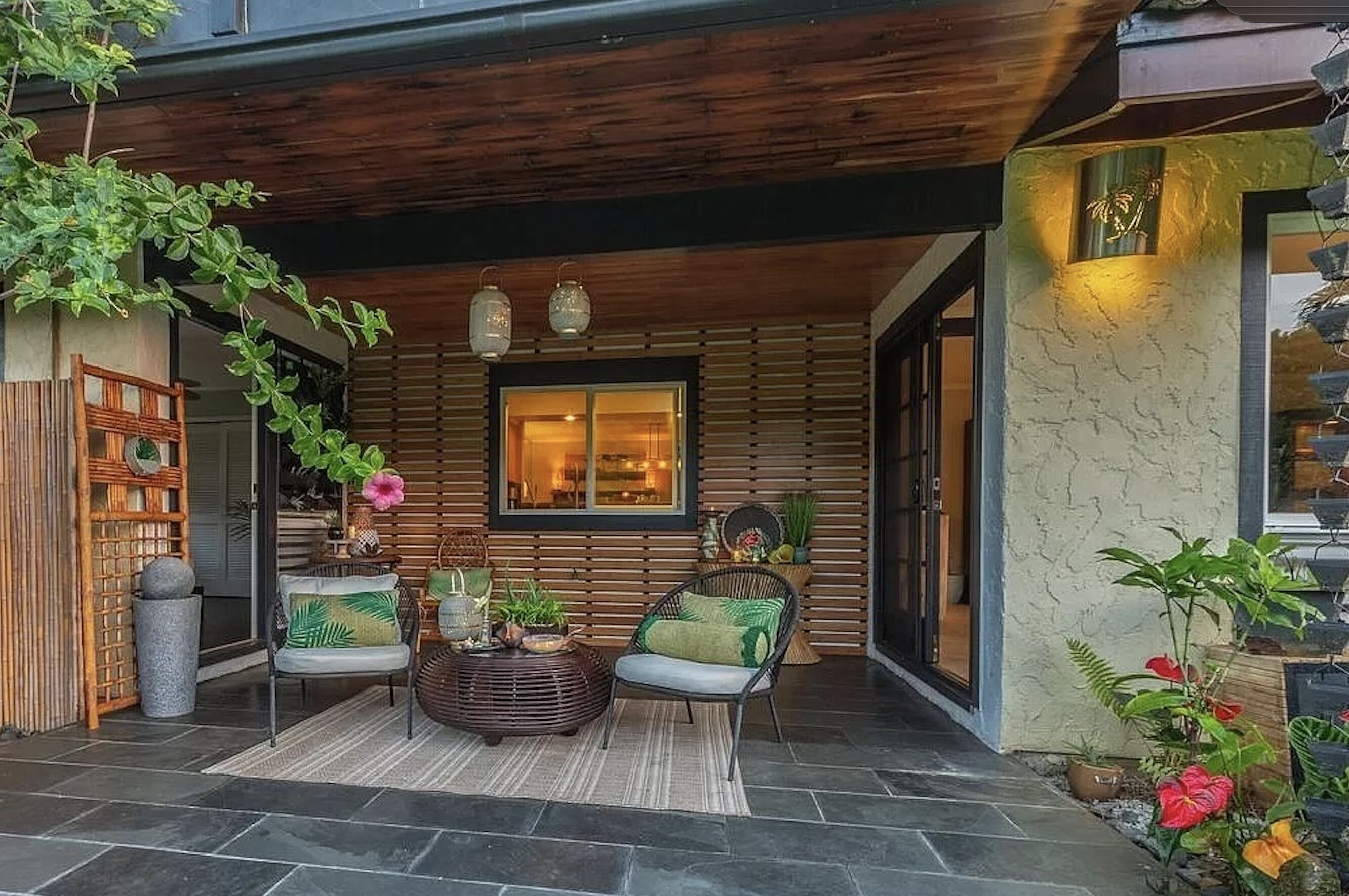 Un espace extérieur accueillant, aménagé avec des meubles en osier et une petite table, entouré de plantes en pot et d’une pergola en bois.