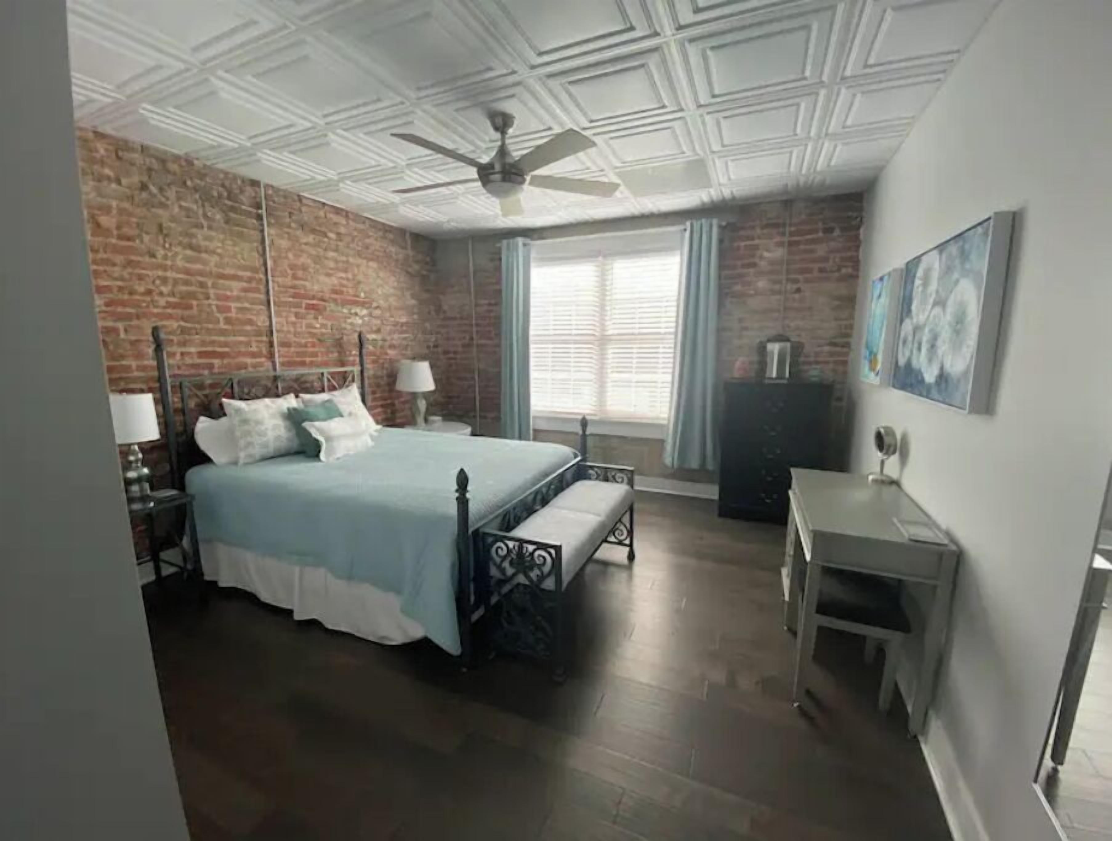 A bedroom with a brick wall, a bed with a canopy, a desk, and a ceiling fan.