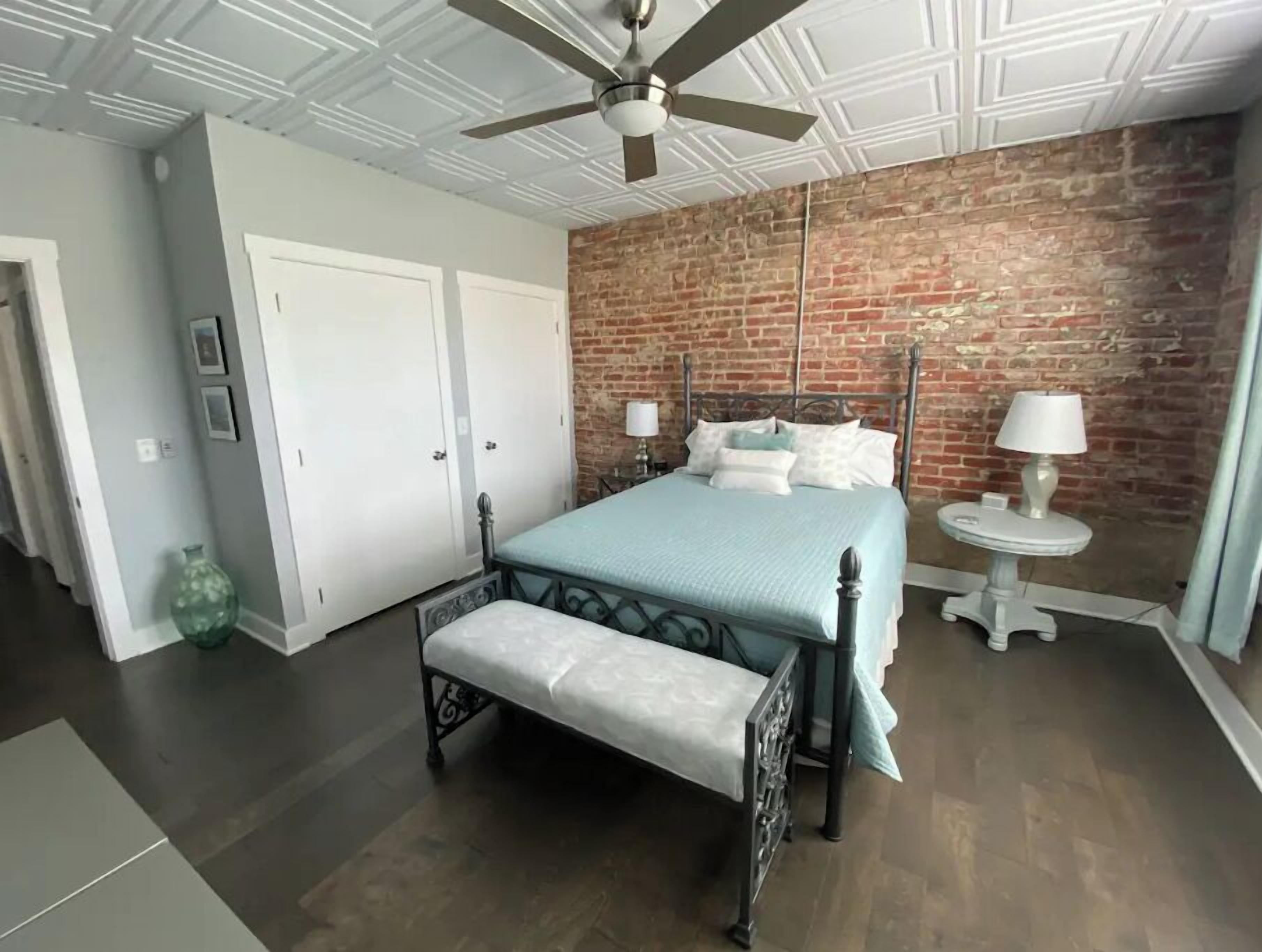 A bedroom with a bed, bench, ceiling fan, brick wall, and a small table with a lamp.