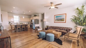 A spacious living room with a brown leather sofa, a wooden coffee table, and blue ottomans. There is a dining area with a wooden table and chairs, and a kitchen in the background. - Ranch House Interior - 4 bedroom, 4 bath home on-site of Galway Downs (Temecula)