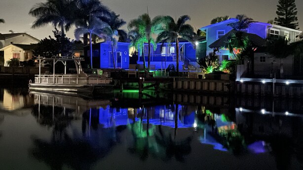 A waterfront property with a dock, palm trees, and illuminated buildings reflecting on the water.