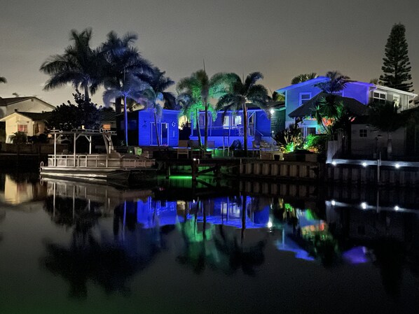 A waterfront property with a dock, palm trees, and illuminated buildings reflecting on the water.