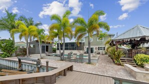 A wooden deck with a pool, palm trees, and a building with a covered area.
