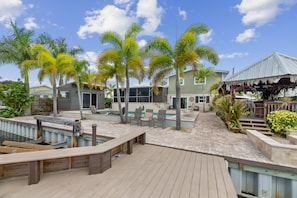 A wooden deck with a pool, palm trees, and a building with a covered area.