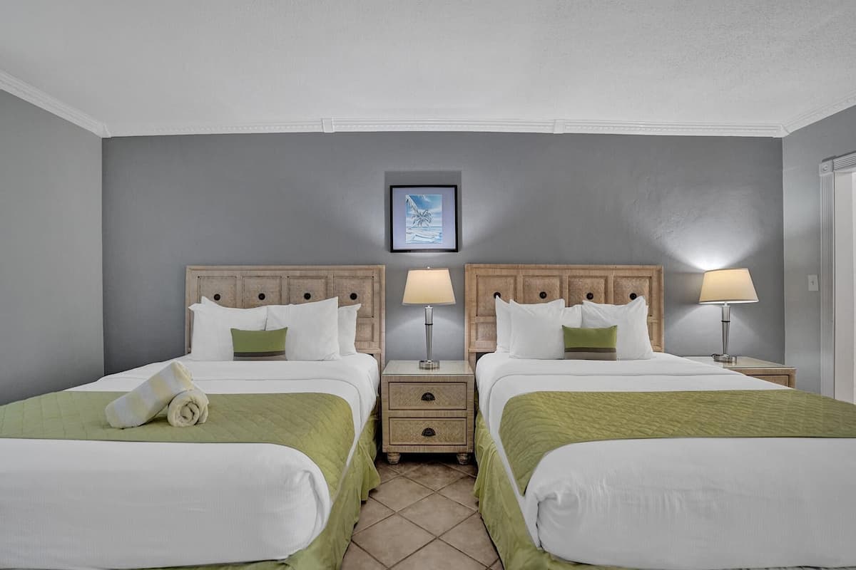 Une chambre d’hôtel avec deux lits, chacun recouvert d’une couvre-lit verte et doté de draps blancs, et flanquée de têtes de lit en bois. Il y a des tables de chevet avec des lampes et un petit tiroir. Un tableau encadré est accroché au mur au-dessus des lits.