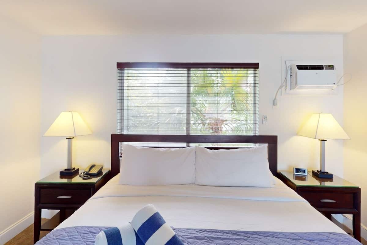 Une chambre d’hôtel comprenant un lit, deux tables de chevet, une fenêtre avec stores, un appareil de climatisation et une petite table de nuit avec un téléphone et une lampe.