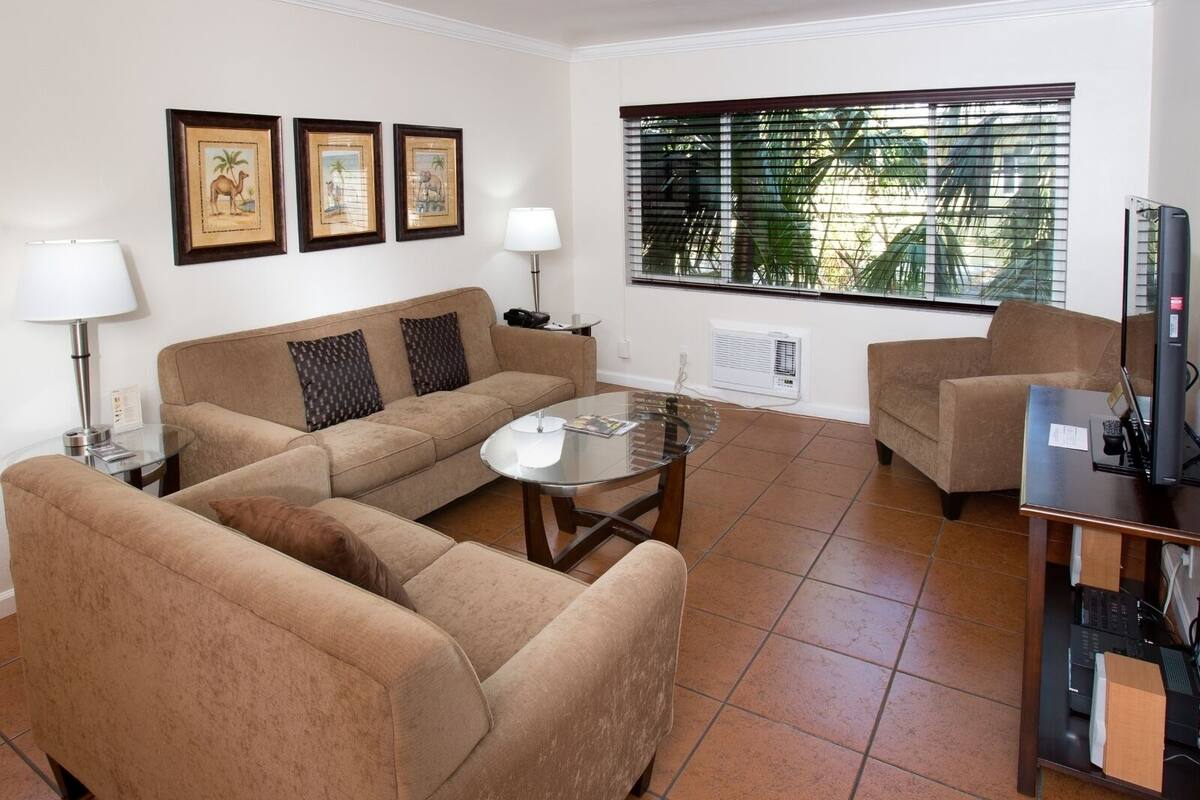 Un salon avec un canapé beige, une table basse en verre, un meuble pour la télévision avec un téléviseur à écran plat et une fenêtre avec des stores.