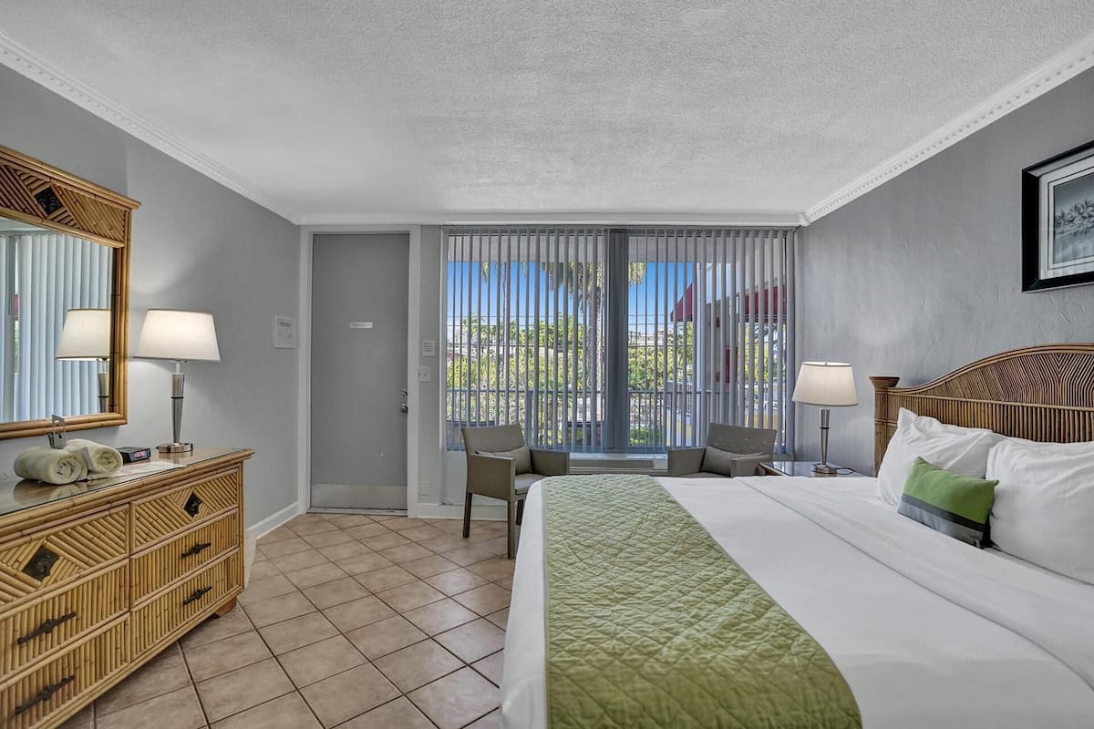 Une chambre d’hôtel avec un lit, une commode, une chaise, une fenêtre avec des stores et une vue sur la verdure.