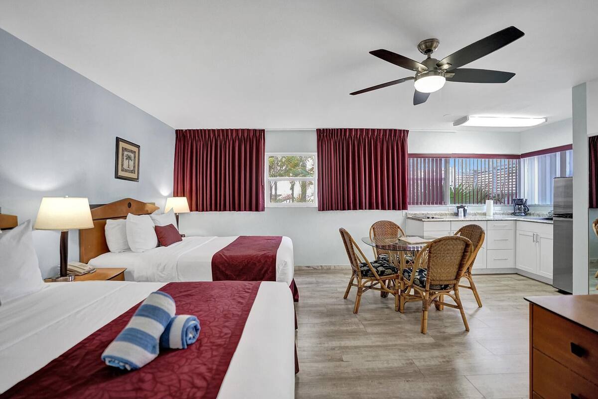 Une chambre d’hôtel avec deux lits, une table à manger avec des chaises, un ventilateur de plafond et un coin cuisine.