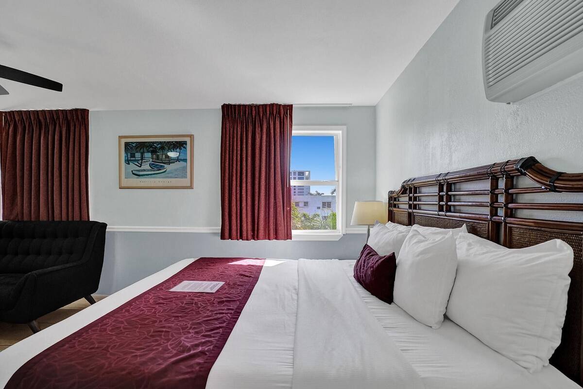 Une chambre d’hôtel avec un lit, une chaise, une fenêtre avec des rideaux, un tableau et un climatiseur fixé au mur.
