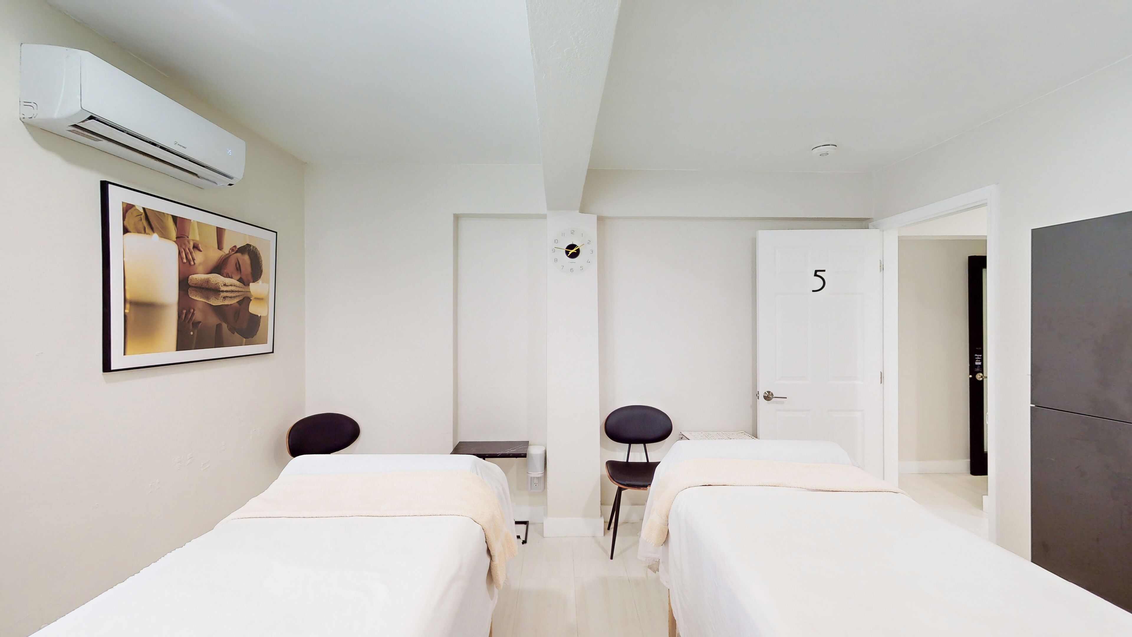 Une chambre d’hôtel avec deux lits simples, un bureau, une chaise, un appareil de climatisation, une horloge et une porte portant le numéro 5.
