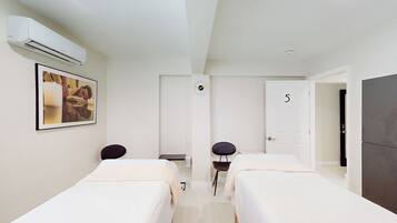 Une chambre d’hôtel avec deux lits simples, un bureau, une chaise, un appareil de climatisation, une horloge et une porte portant le numéro 5.