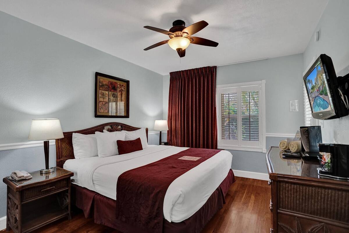 Une chambre d’hôtel avec un lit, deux tables de chevet, un ventilateur de plafond, une fenêtre avec des rideaux, une télévision et un tableau au mur.