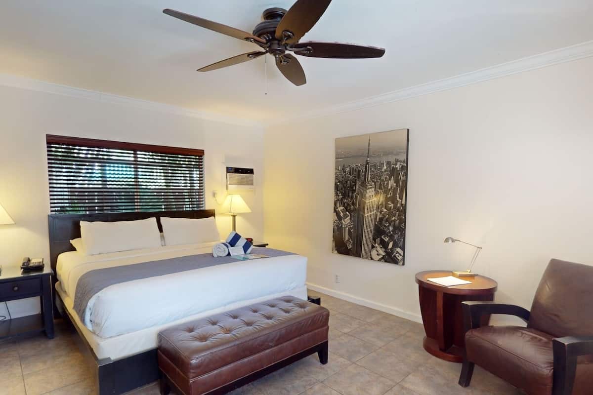 Une chambre d’hôtel comprenant un lit, un ventilateur de plafond, une fenêtre avec stores, une table de chevet avec une lampe, une chaise et un climatiseur fixé au mur.