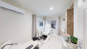 Quarto moderno e bem iluminado com uma cadeira de massagem, uma pequena mesa com uma planta e uma placa indicando serviços de massagem.