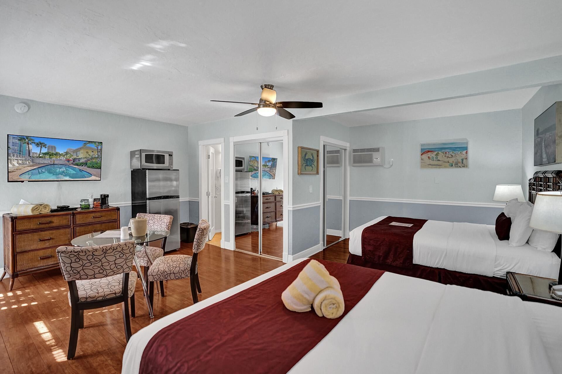 A hotel room with a bed, a desk with a computer, a chair, a microwave, a refrigerator, and a ceiling fan.