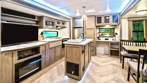 Cocina moderna para vehículo recreativo con electrodomésticos integrados, comedor y un amplio ventanal con vistas a la vegetación.