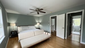 Un dormitorio con cama, lámparas de noche, ventilador de techo y vista a un baño por una puerta abierta.