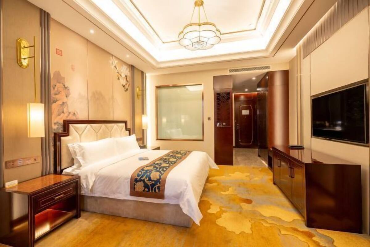 A hotel room with a bed, bedside table, wall art, a large window, and a TV.