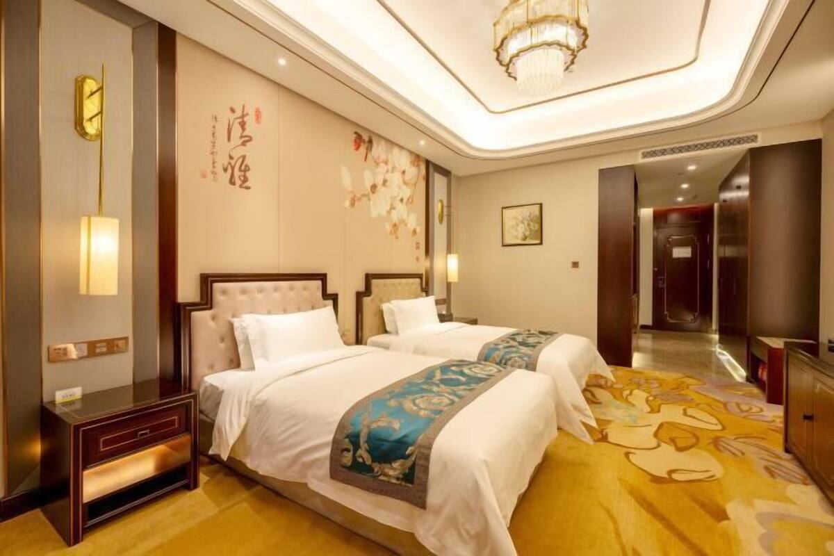 A hotel room with two beds, a chandelier, a wall clock, and a decorative wall piece.