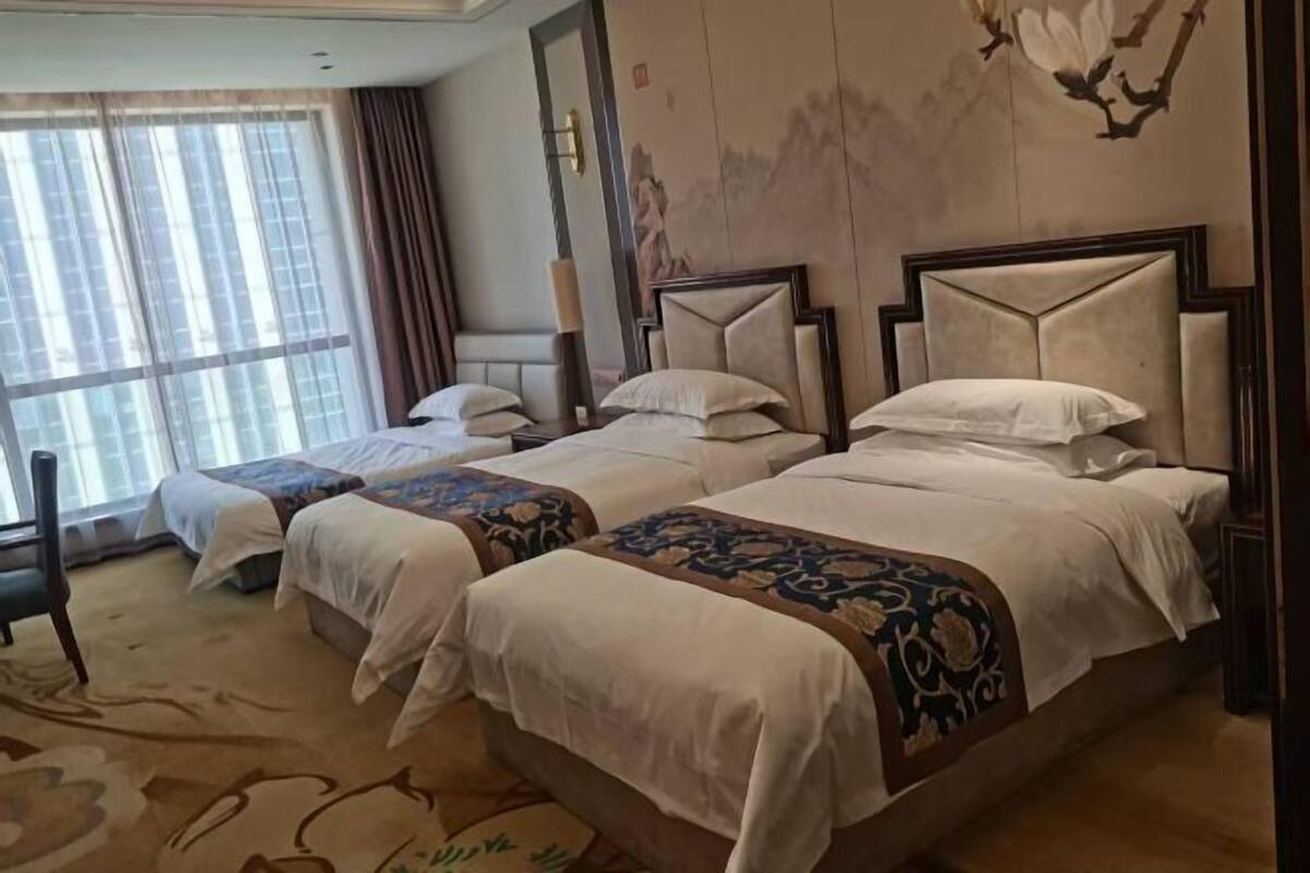 A hotel room with two beds, a large window with curtains, a decorative wall painting, and a carpet with a floral pattern.