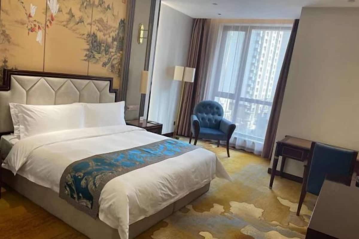 A hotel room with a bed, a chair, a desk, a lamp, and a view of buildings through the window.