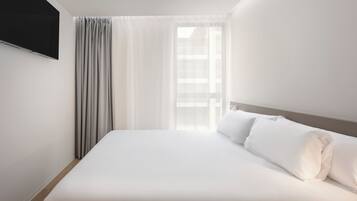 A hotel room with a bed, curtains, a TV mounted on the wall, and a window with sheer curtains.