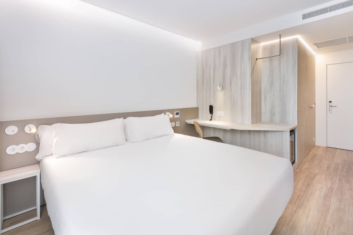 A modern hotel room with a large bed, a bedside table, a built-in desk, and a mirror.