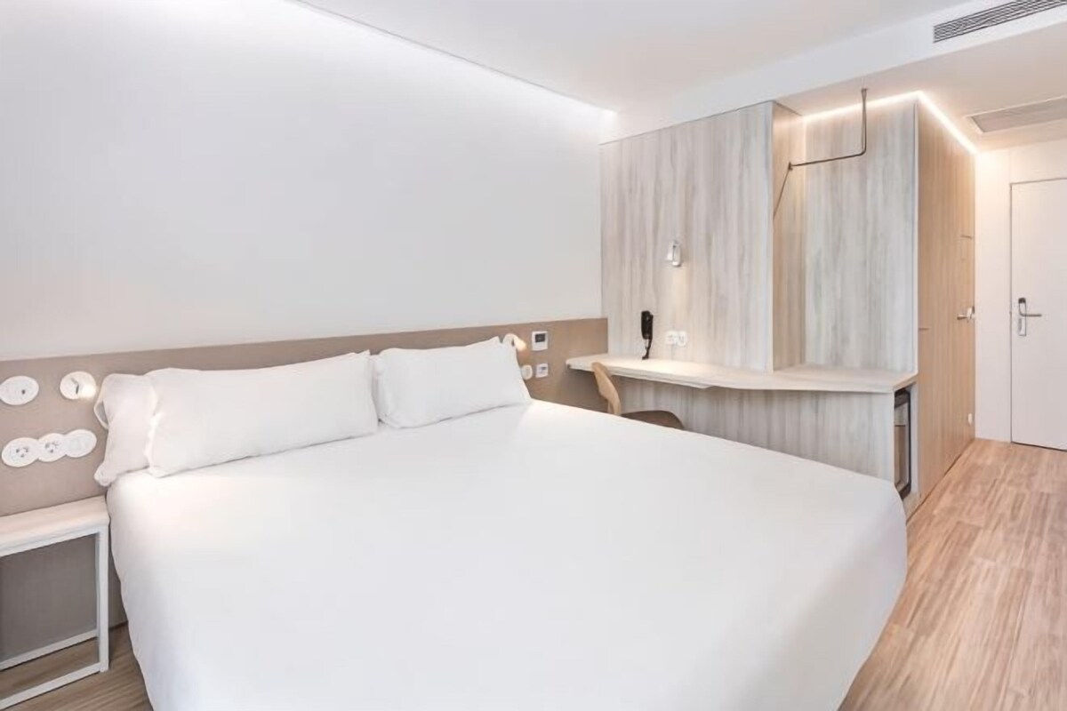 A modern hotel room with a large bed, bedside table, and a built-in vanity with mirror and lighting.