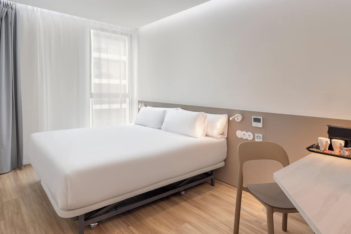 A neatly arranged hotel room with a bed, a chair, a small table, and a window with curtains.