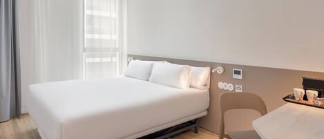 Habitación de hotel ordenada con cama, una silla, una mesita y ventana con cortinas.