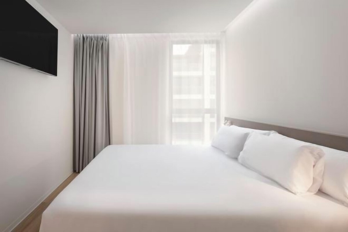 A hotel room with a large bed, a flat-screen TV mounted on the wall, and a window with sheer curtains.
