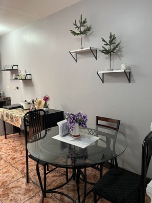 Een eetgedeelte met een ronde glazen tafel, zwarte stoelen, een vaas met paarse bloemen en twee wandplanken met kleine potplanten.