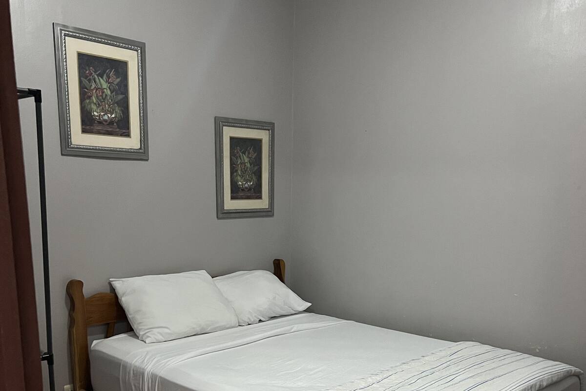 Een enkel bed met houten frame en witte beddengoed in een kamer met lichtgrijze wanden en bruine betegelde vloer.