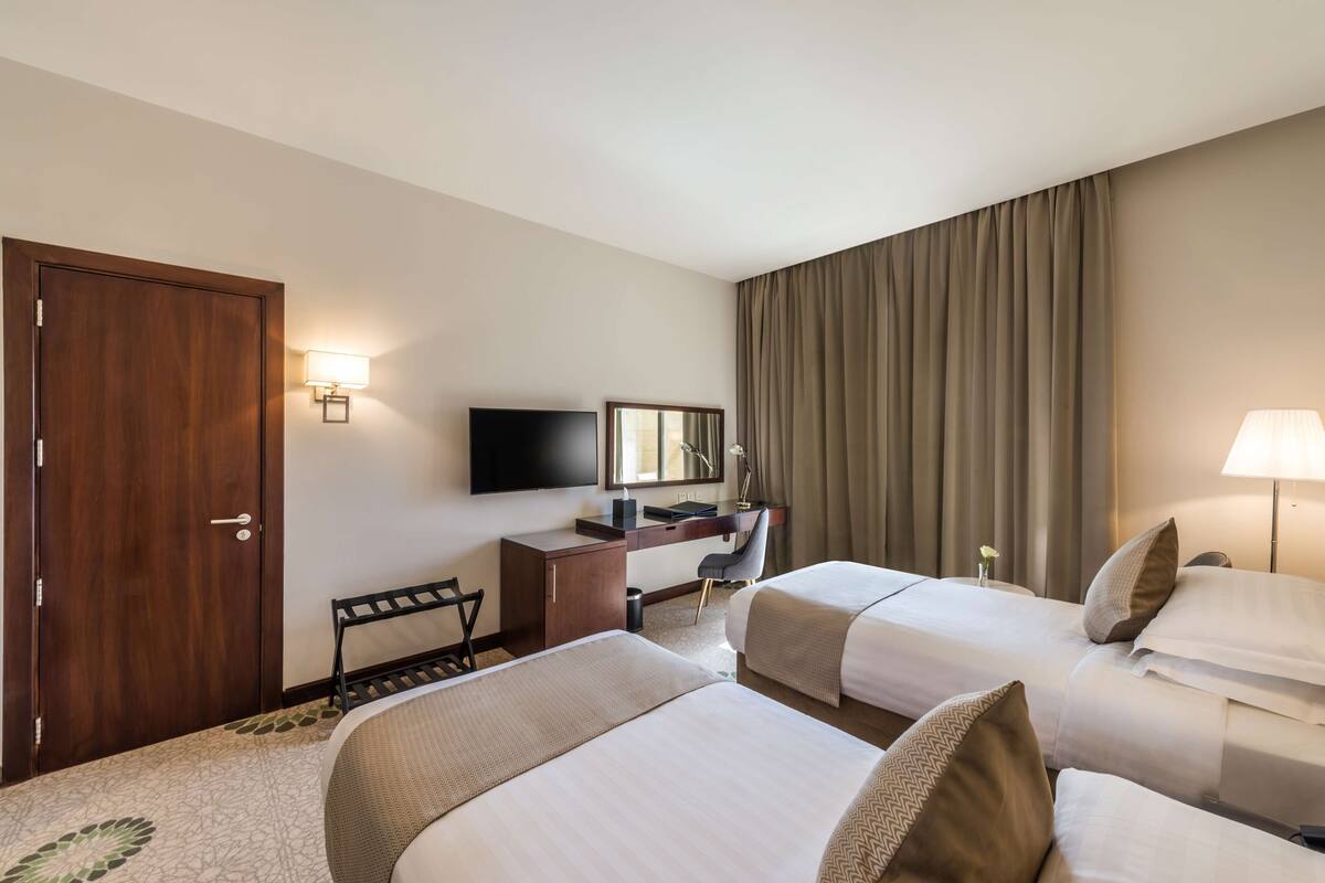 Ein Hotelzimmer mit zwei Betten, einem Schreibtisch, einem Flachbildfernseher, einem Sessel, einer Schreibtischlampe und einem großen Fenster mit Vorhängen.