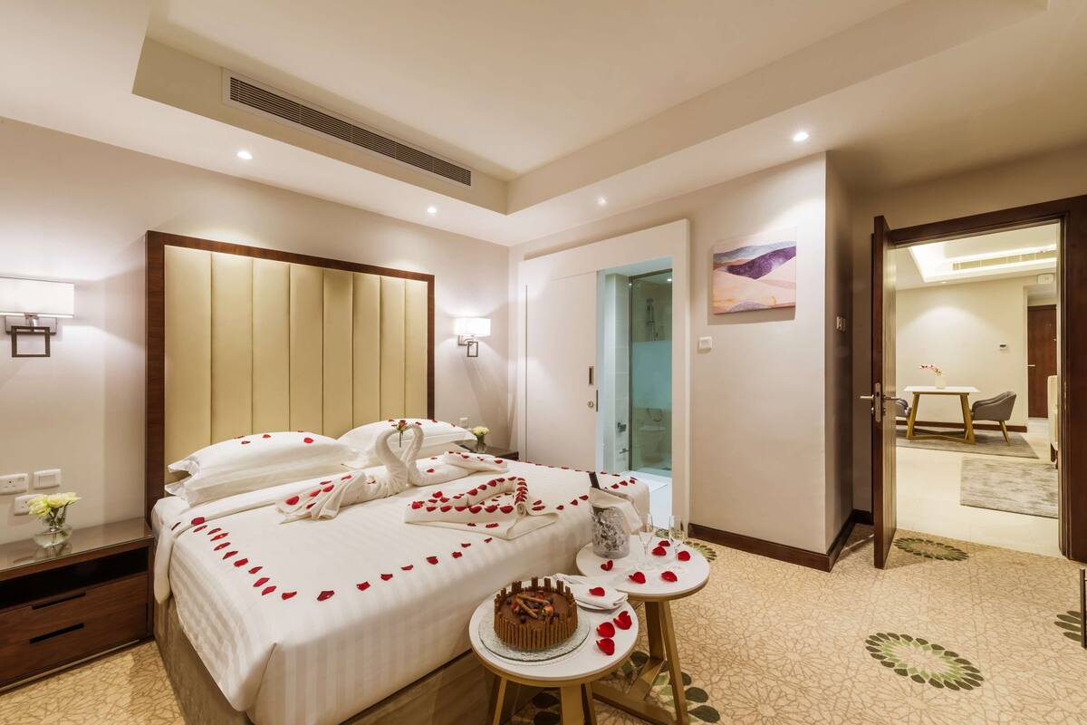 Ein Hotelzimmer mit einem großen Bett, einem Nachttisch mit Kuchen und einem sichtbaren Duschbereich hinter einer Glastür.