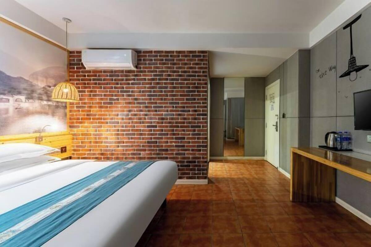 Una habitación de hotel con una pared de acento de ladrillo, una cama con colcha azul y blanca a rayas, un banco de madera, un televisor y un aire acondicionado instalado en la pared.