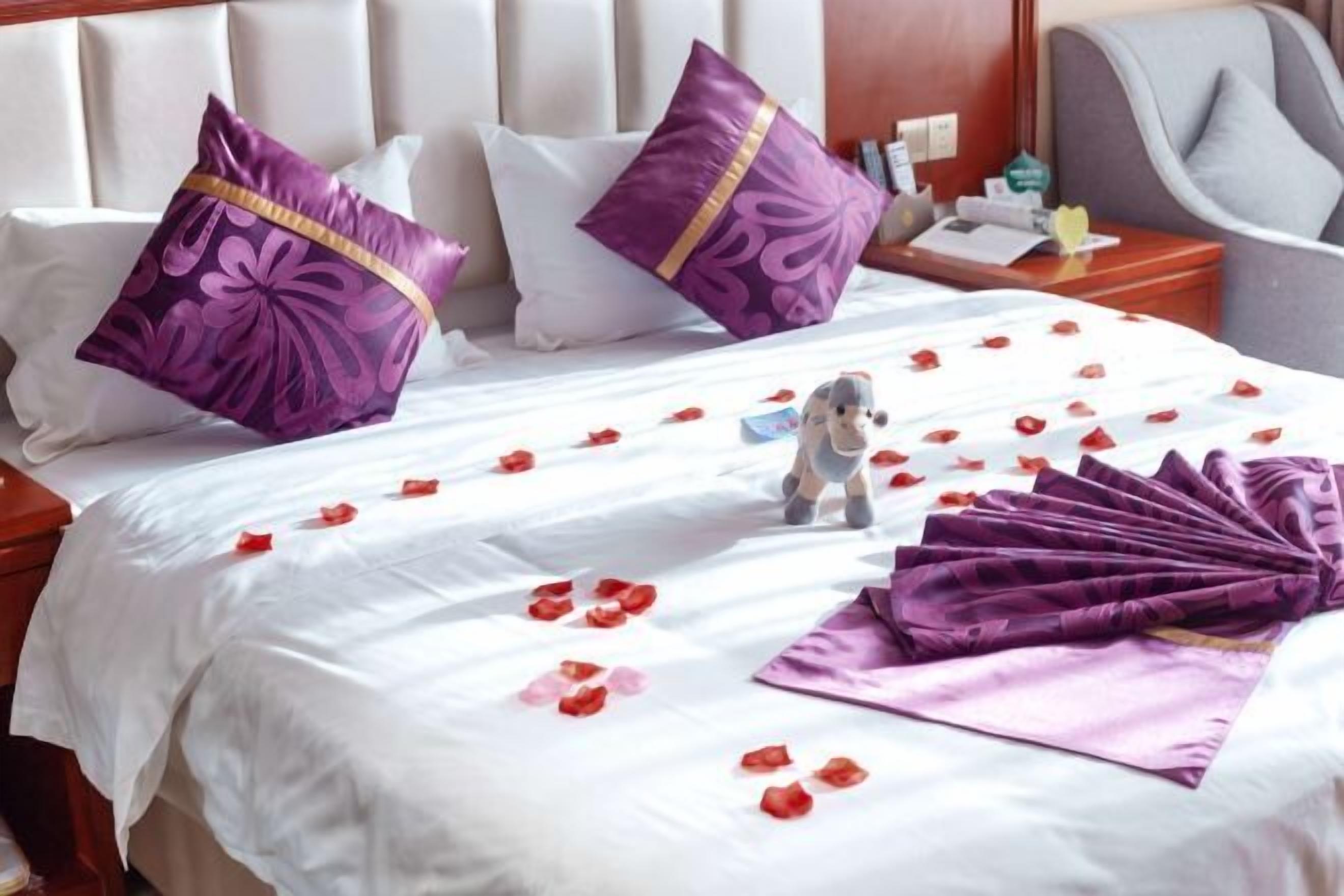 Ein Hotelzimmer mit einem Bett, zwei violetten Kissen, einer kleinen Figur und Rosenblüten auf dem Bett.