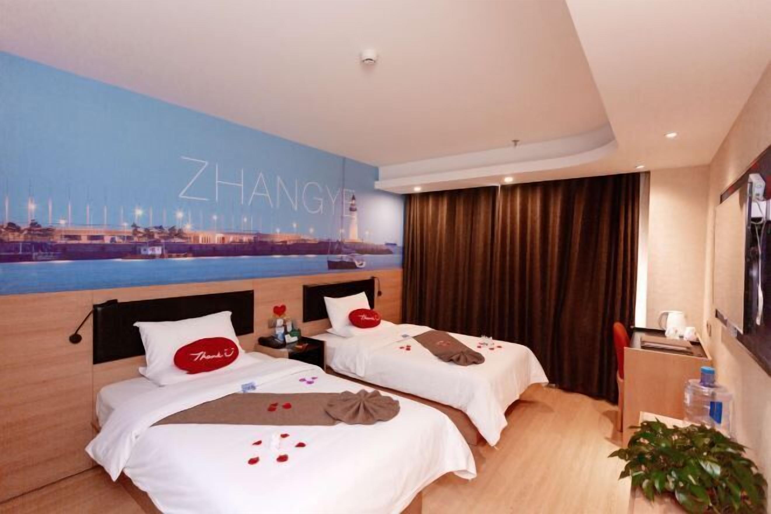 A hotel room with two beds, a cityscape mural, a view of the water, and a plant on the side.