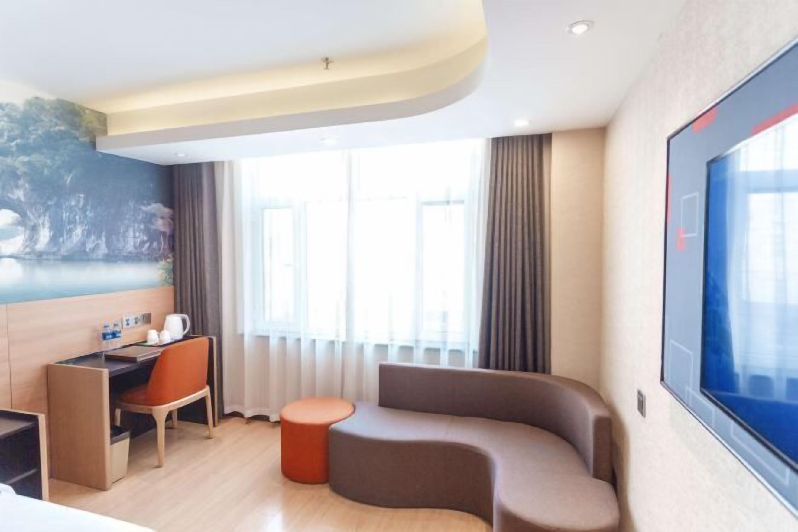 Habitació d'hotel moderna amb un sofà corbat, un otomanet taronja petit, un escriptori amb una cadira i una finestra gran amb cortines.