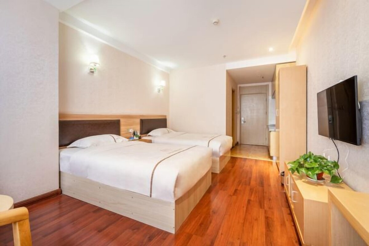 Ein Hotelzimmer mit zwei Betten, Holzboden, einem an der Wand montierten Fernseher und einer kleinen Pflanze auf einem Nachttisch.