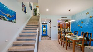 Una escalera conduce a un comedor con un muro azul con motivos marinos y vista al océano a través de una ventana.