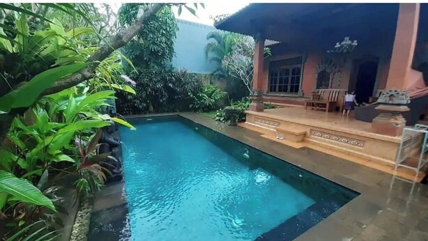 Ein rechteckiger Pool mit Fliesenrand, umgeben von üppiger Bepflanzung und einem Gebäude mit einem geflasten Dach und einem Balkon.