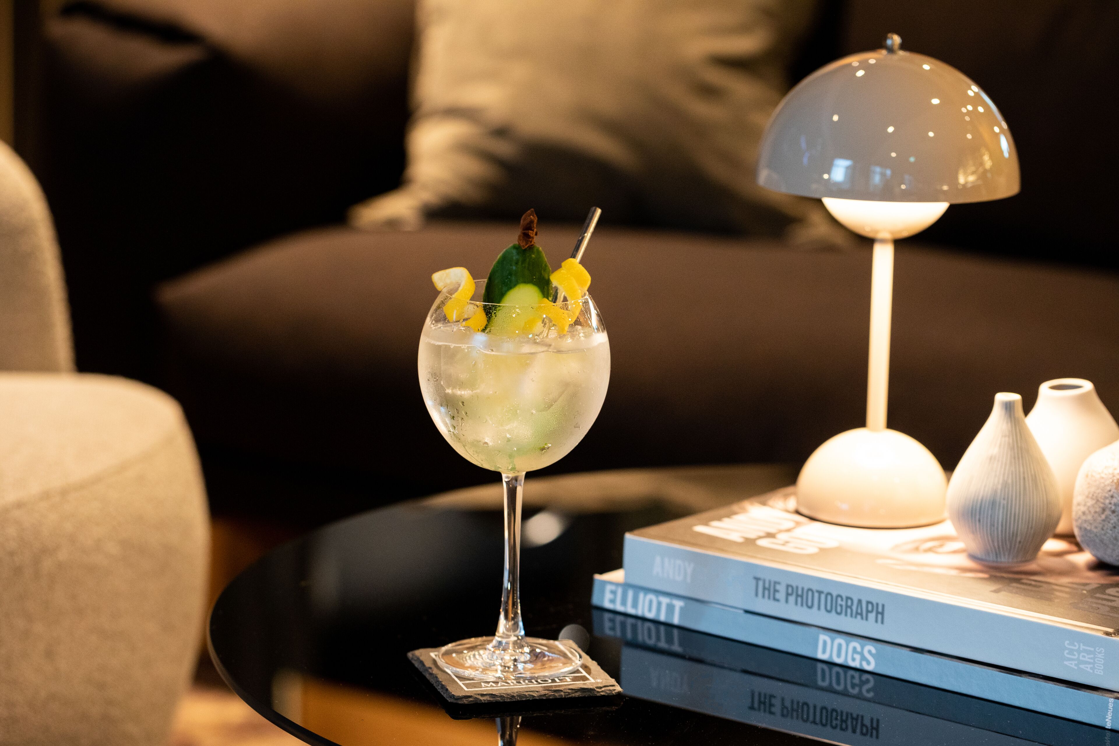 A cocktail glass with a lime wedge and a straw on a coaster, placed on a black table beside a stack of books titled 'The Photograph' and 'Dogs'.